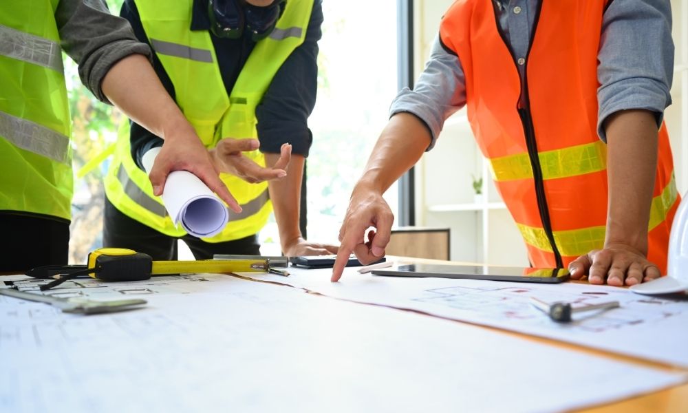 dwie osoby w kamizelce ż&oacute;łtej oraz pomarańczowej sprawdzające papiery oraz rysunki architektoniczne